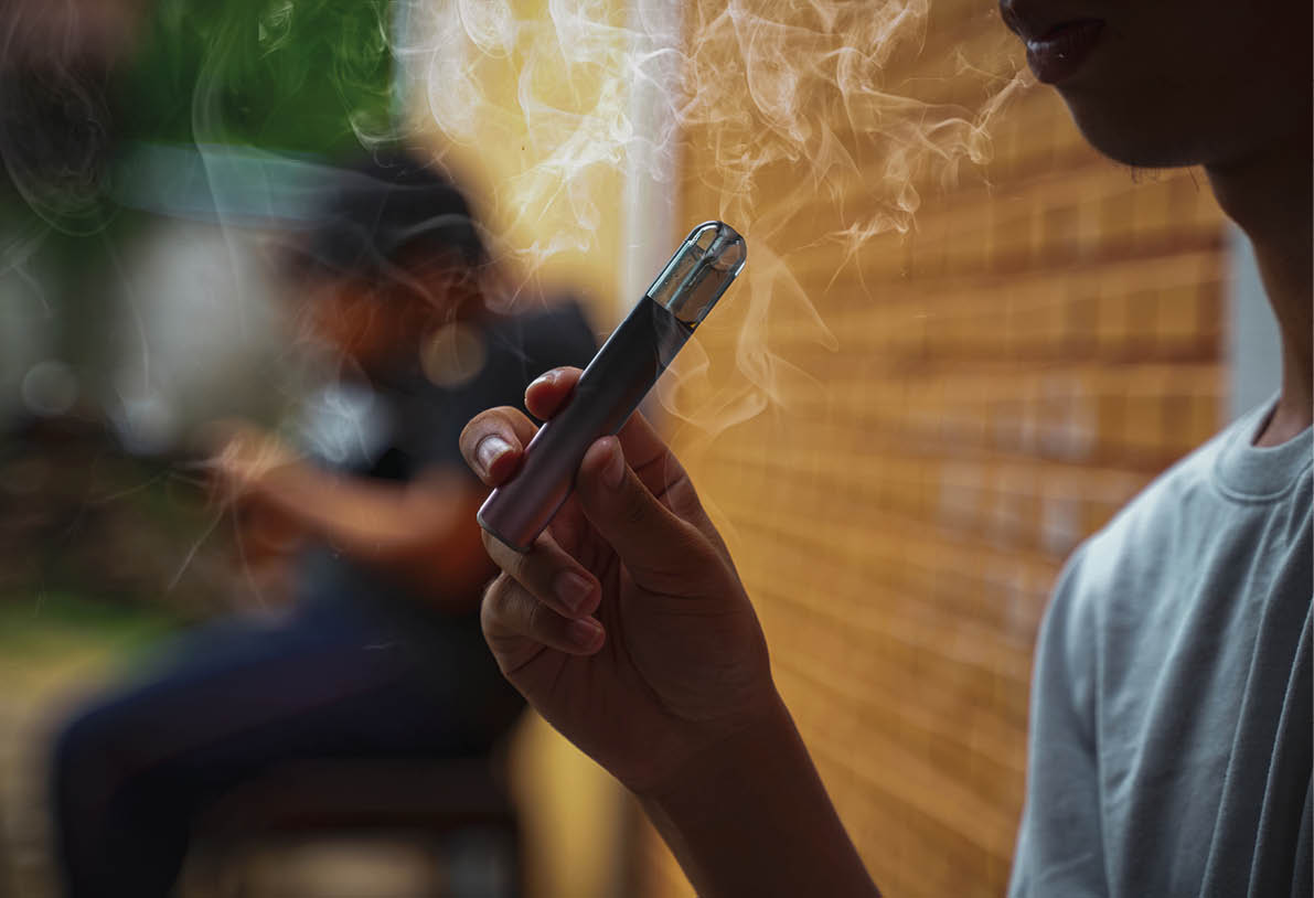 Asian male teenager holds a smoked e-cigarette in his hand while sitting during recess in a hidden corner with his friends at school.
