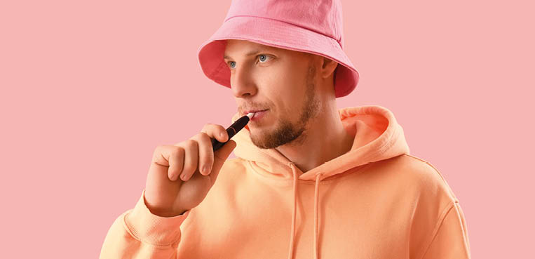 Young man in bucket hat smoking disposable electronic cigarette on pink background