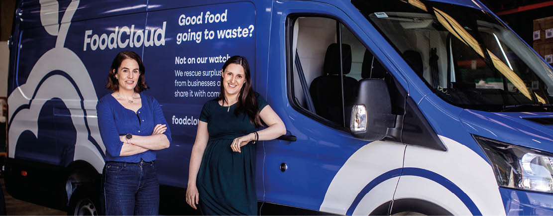Two women standing in front of a blue and white van. Description generated by AI