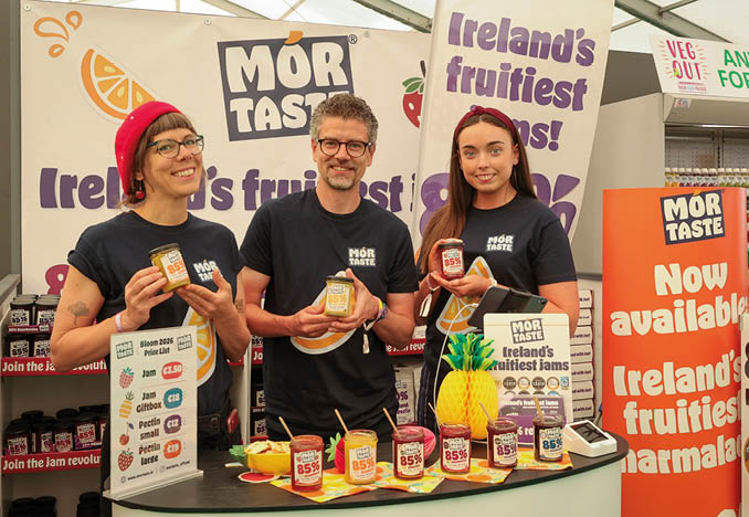 Three people are standing in front of a table with jars of jam. Description generated by AI
