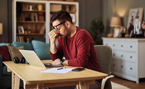 A tired and overworked freelance man with glasses sitting at a desk at home in front of a laptop and pinching his nose.