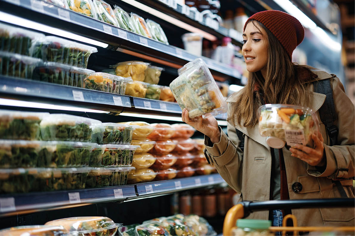 Smiling woman reading label on food package while buying groceries from refrigerated section in supermarket.