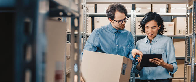 Female Inventory Manager Shows Digital Tablet Information to a Worker Holding Cardboard Box, They Talk and Do Work. In the Background Stock of Parcels with Products Ready for Shipment.