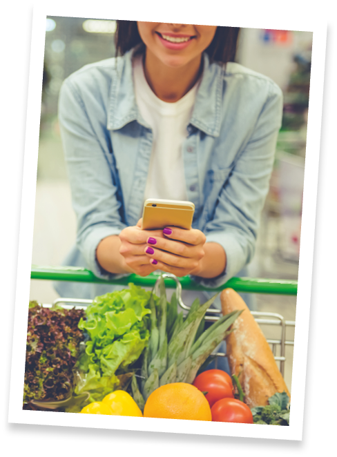 Couple in the supermarket. Cropped image of girl leaning on shopping cart, using a mobile phone and smiling, in the background her boyfriend is choosing food