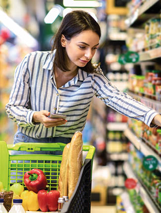 Portrait Of Millennial Lady Holding And Using Smartphone Buying Food Groceries Walking In Supermarket With Trolley Cart. Female Customer Shopping With Checklist, Taking Products From Shelf At The Shop