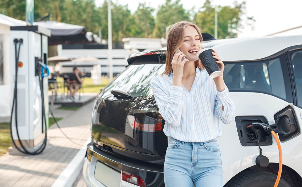 Happy young adult woman smiling wide, looking away, charging automobile battery from small public station, standing near electric car, drinking coffee and talking on smartphone