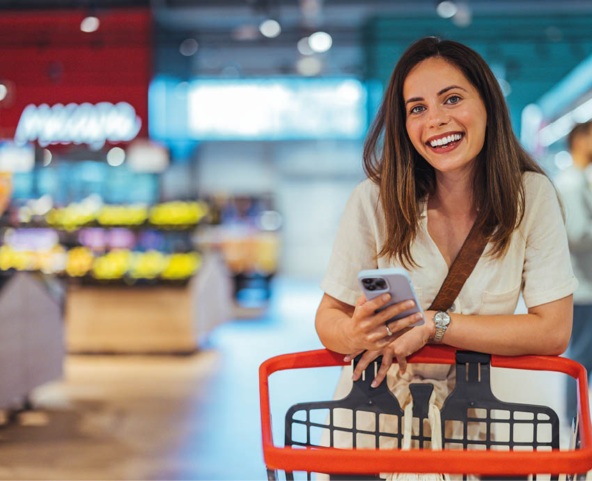 Portrait of happy woman buying groceries in supermarket and looking at camera. Purchasing Goods with Smartphone at Grocery Store. Female customer shopping with smartphone checklist