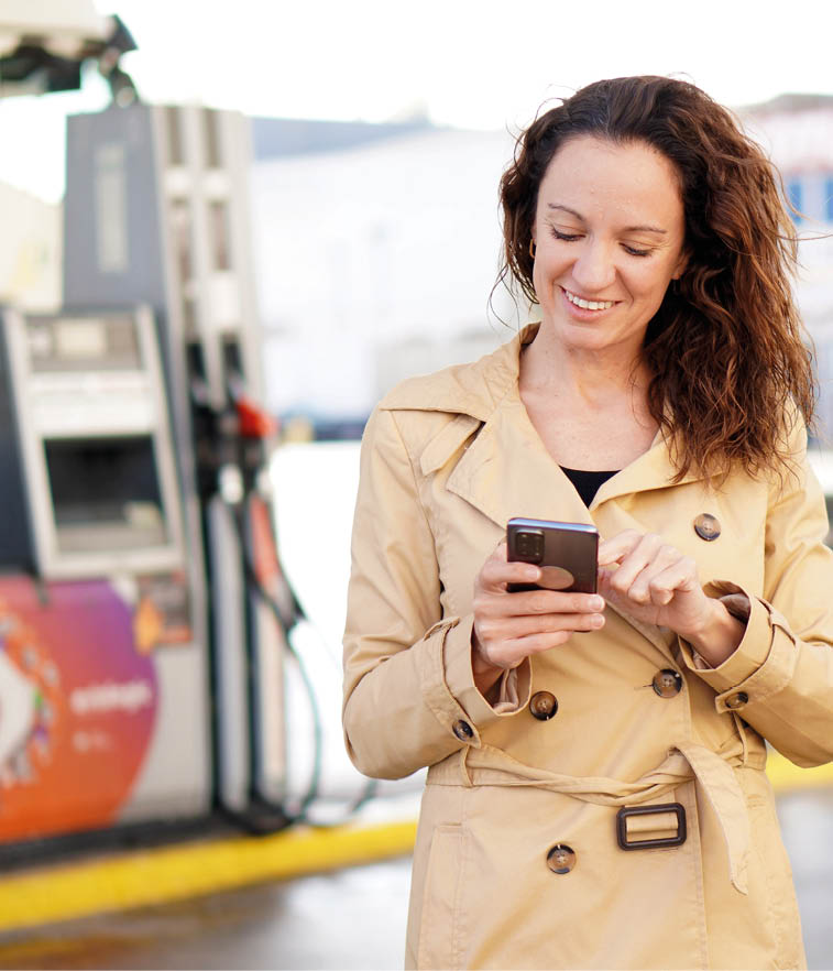 A middle-aged woman with curly hair and a trench coat smiles while using her cell phone with an out-of-focus gas station in the background.