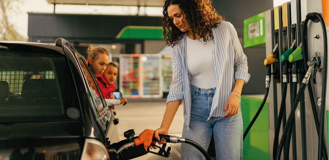 A young mother fills up gas tank at a gas station while her daughters look out the car window