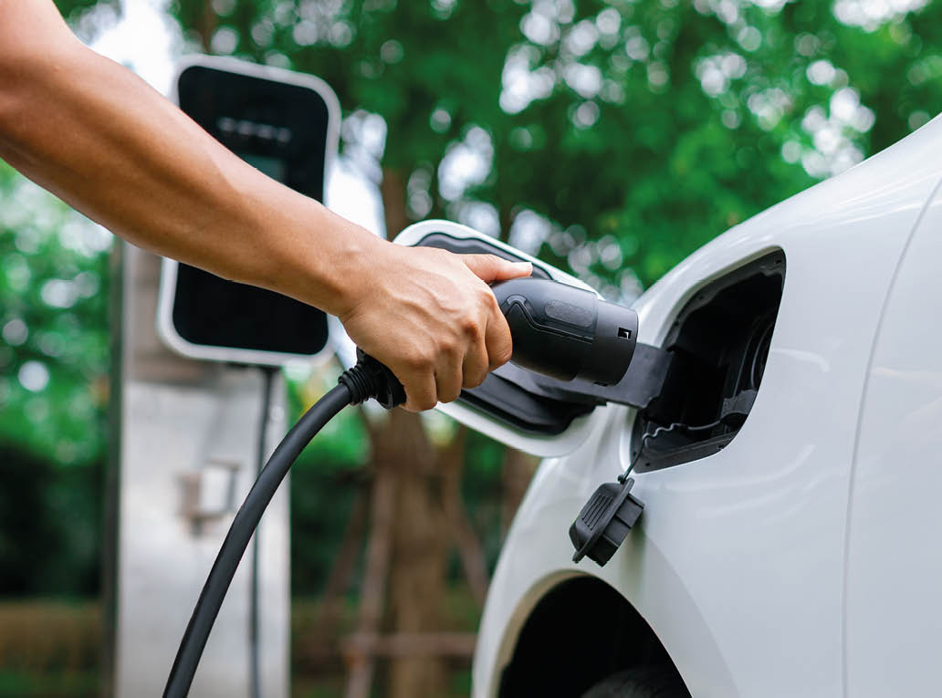 Hand inserting EV charging plug to electric vehicle in focus shot with blurred background of outdoor natural greenery. Progressive sustainable energy powered electric charging station for rechargeable
